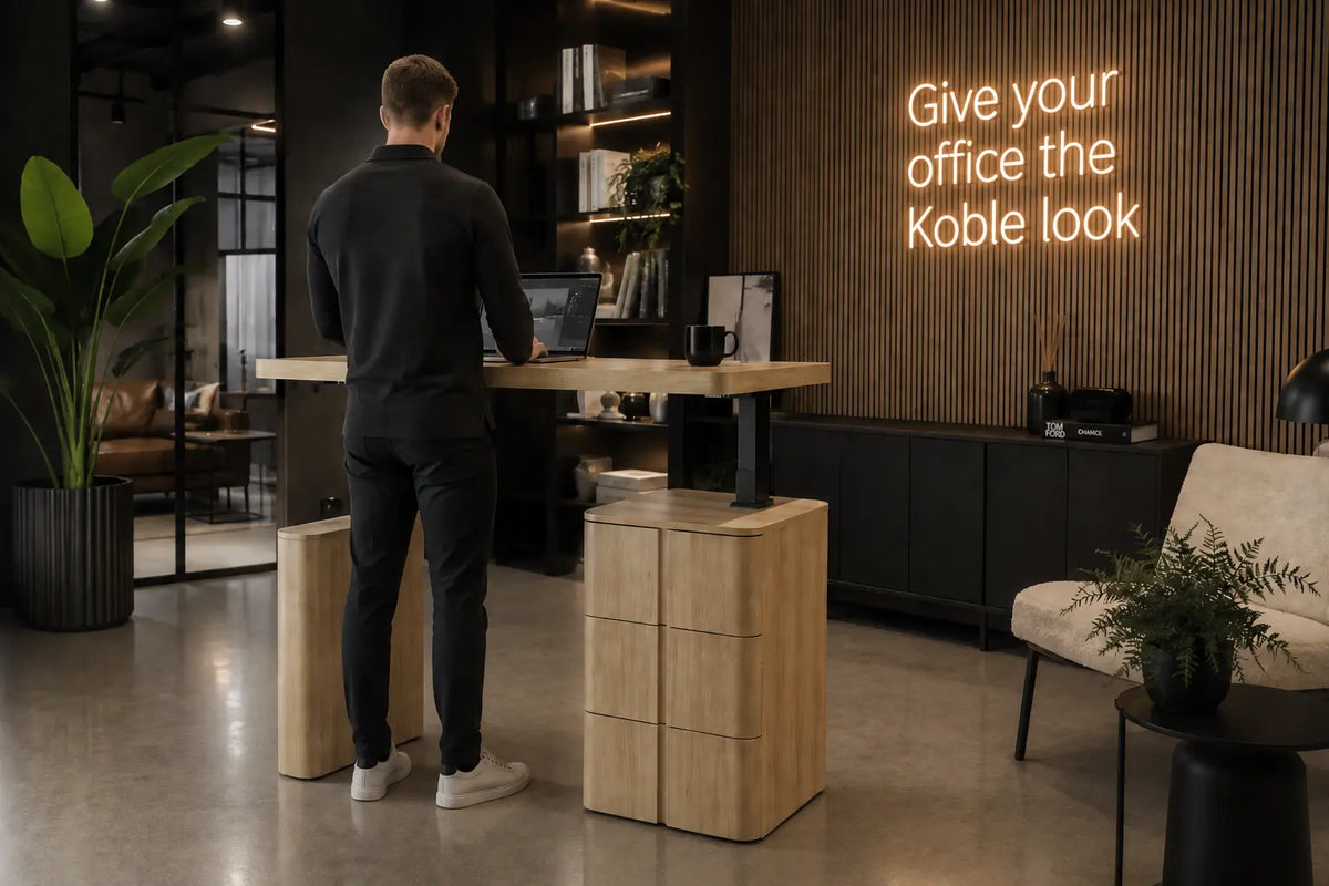 A man stands at a modern standing desk in a contemporary office space. The desk has a light natural wood top and drawer base with black metal legs.